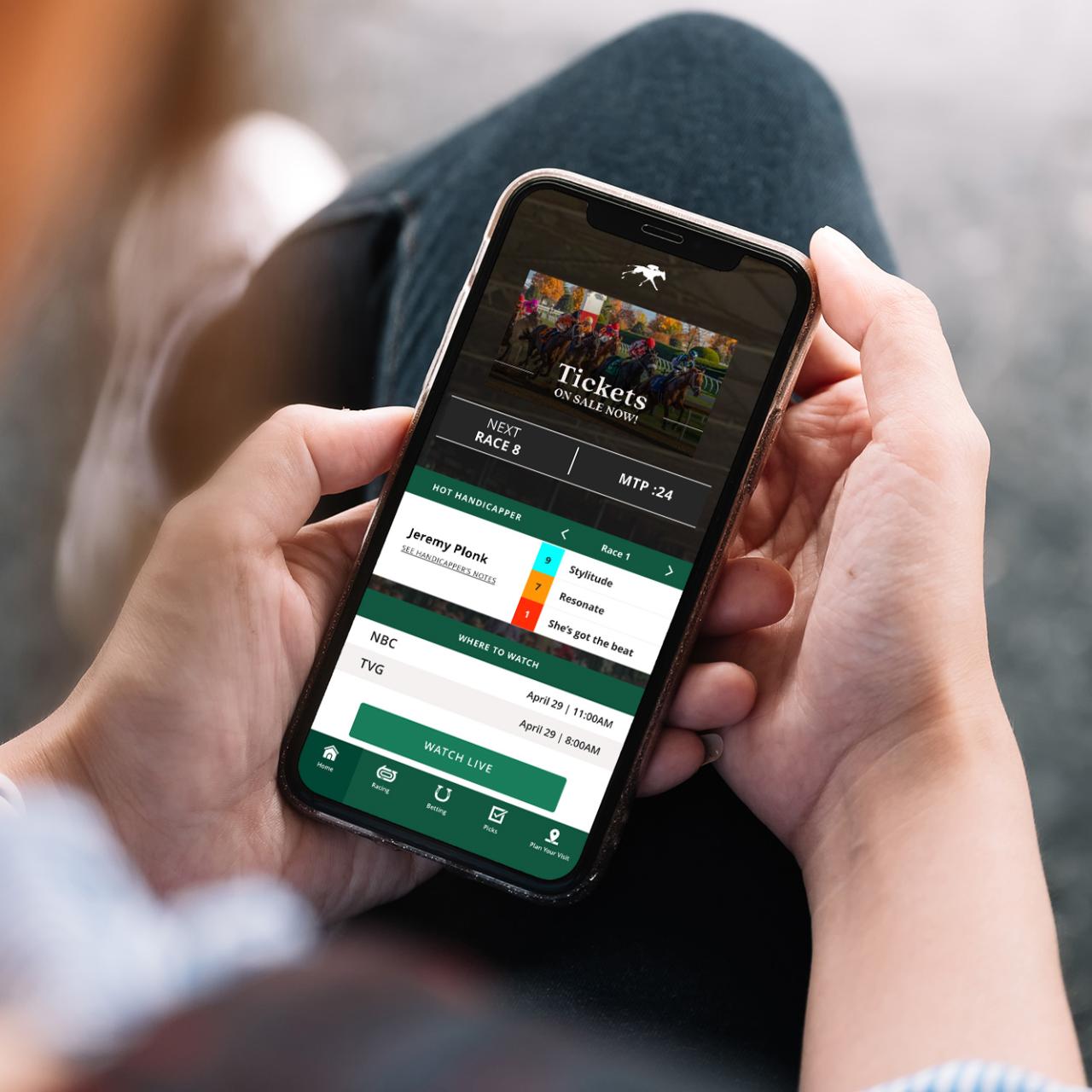 Hands holding a phone showing Keeneland App-Home screen. This new feature has the hottest handicappers, watch live, and your favorites horse picks per race.